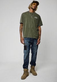 T-shirt vert olive avec logo, jean bleu avec des patches noirs, et bottes beige. Les accessoires incluent des lunettes de soleil jaunes et des colliers superposés.