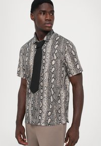 Chemise à manches courtes avec un motif imprimé serpent gris et noir, dotée d'un col et d'une cravate noire, portée avec un pantalon beige. Texture lisse.