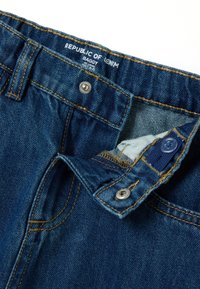 Jeans holgados en denim oscuro con costuras amarillas, cierre de botón plateado y forro interior azul claro. La etiqueta dice "República del Denim."