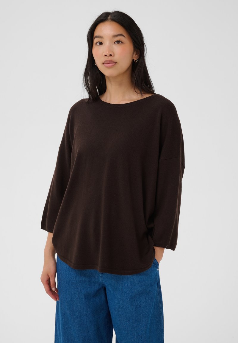 Femme aux longs cheveux noirs portant un pull marron foncé ample et un jean bleu, debout devant un fond clair uni.