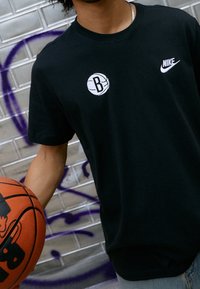 Camiseta de algodón negra con un gráfico blanco de baloncesto que presenta un logo de "B" y un pequeño logo de Nike blanco en el pecho, acompañada de un balón de baloncesto.