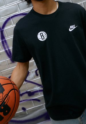 Camiseta de algodón negra con un gráfico blanco de baloncesto que presenta un logo de "B" y un pequeño logo de Nike blanco en el pecho, acompañada de un balón de baloncesto.