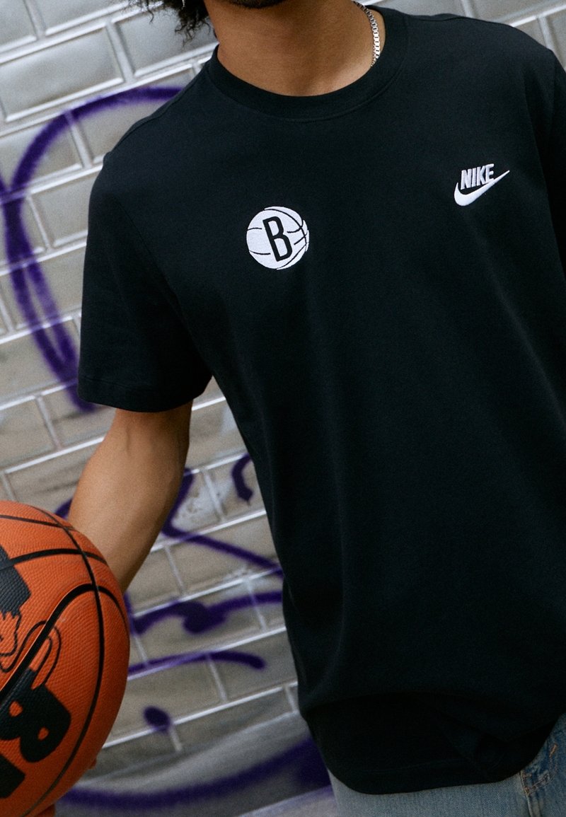 Camiseta de algodón negra con un gráfico blanco de baloncesto que presenta un logo de "B" y un pequeño logo de Nike blanco en el pecho, acompañada de un balón de baloncesto.