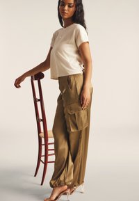 Camiseta de crema recortada con mangas cortas combinada con pantalones cargo de satén verde oliva con bolsillos y puños elásticos. Silla de madera roja al fondo.