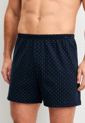 Uomo che indossa boxer blu scuro con motivo a piccoli puntini bianchi, visibile dalla vita a metà coscia su sfondo uniforme.