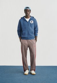 Μπλε φούτερ με φερμουάρ και λογότυπο, λευκό μπλουζάκι, μπεζ και κόκκινο καρό αθλητικό παντελόνι, και λευκά παπούτσια με ταμπά σόλες. Ανοιχτό μπλε καπέλο.