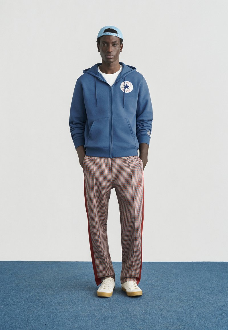 Blå hættetrøje med lynlås og logo, hvid t-shirt, ternede beige og røde løbebukser samt hvide sko med tanfarvede såler. Lyseblå kasket.