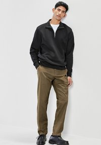 Sweat-shirt zippé noir et pantalon vert olive, associés à des chaussures noires. Textures lisses et couleurs unies, détails de design minimaux.