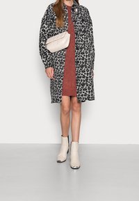 Femme portant un long manteau gris à imprimé léopard sur une robe couleur rouille, avec un sac à bandoulière blanc matelassé et des bottines blanches à bouts argentés.