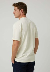 Camiseta de manga corta en color crema, confeccionada con algodón suave, con cuello redondo y un ajuste relajado. La vista trasera muestra un detalle mínimo de costuras.