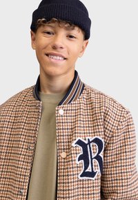 Brown and beige houndstooth patterned jacket, navy striped collar, beige shirt underneath, featuring a blue embroidered "R" on the left chest.