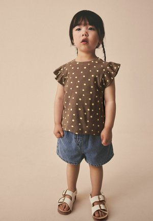 Ung pige med fletninger iført en brun top med polkaprikker, denimshorts og hvide sandaler, stående mod en ensfarvet beige baggrund.