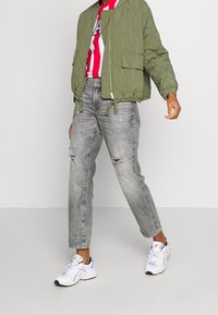 Chaqueta bomber acolchada verde oliva con puños elásticos, camiseta a rayas blanca y roja, vaqueros grises desgastados y zapatillas deportivas blancas.