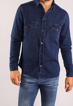 Homme portant une chemise en denim bleu foncé à boutons avec deux poches poitrine et un jean bleu délavé moyen sur fond uni.