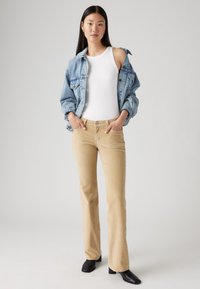 Jeansjakke i lys blå, hvit tanktop og beige kordfløyelsbukser med vid i bena. Modellen står med hendene i lommene, svarte ankelboots.