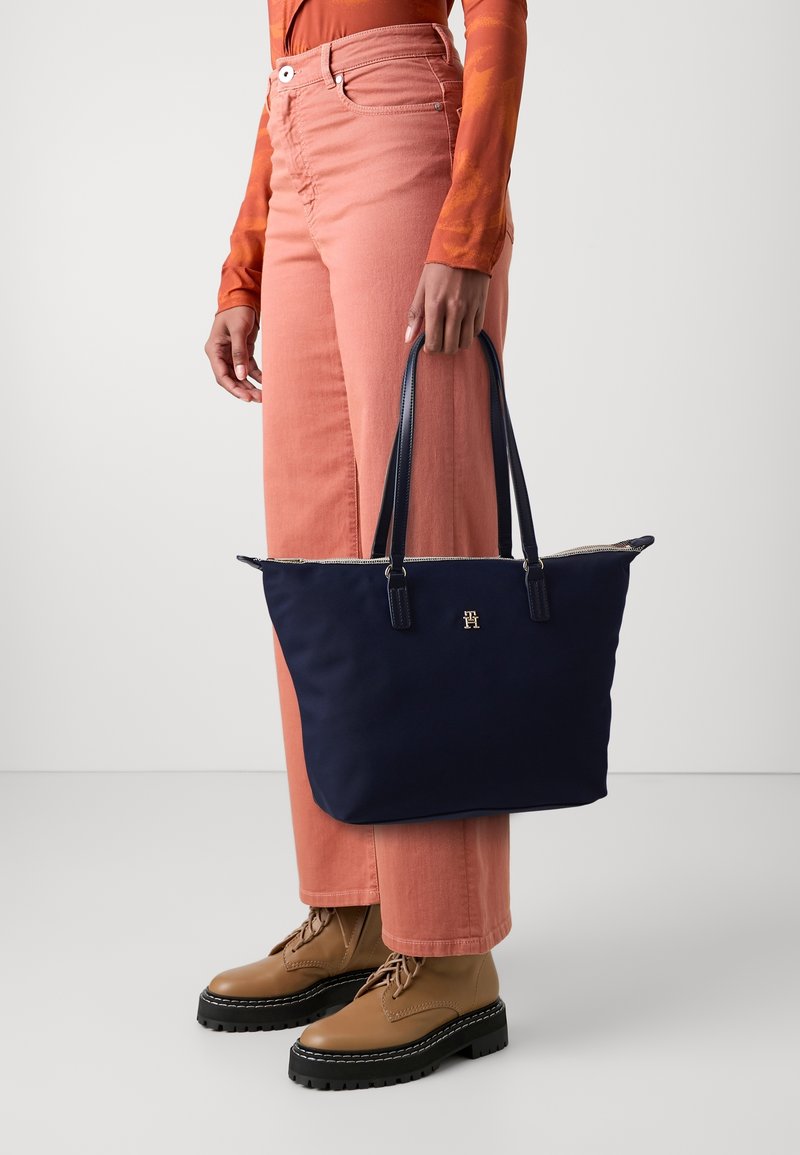 Personne portant un haut orange à manches longues et un pantalon rose, tenant un sac cabas bleu marine avec double poignées et un petit logo doré.