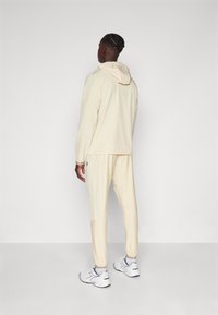 Lys beige treningsdrakt med hette, tapered bukser, subtil tekstur og elastiske mansjetter. Bært med hvite joggesko med blå detaljer.