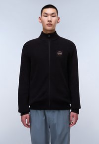 Sweater in maglia nera con zip, colletto alto, texture a coste e una patch con logo sul petto. Modello che indossa pantaloni grigi chiari.