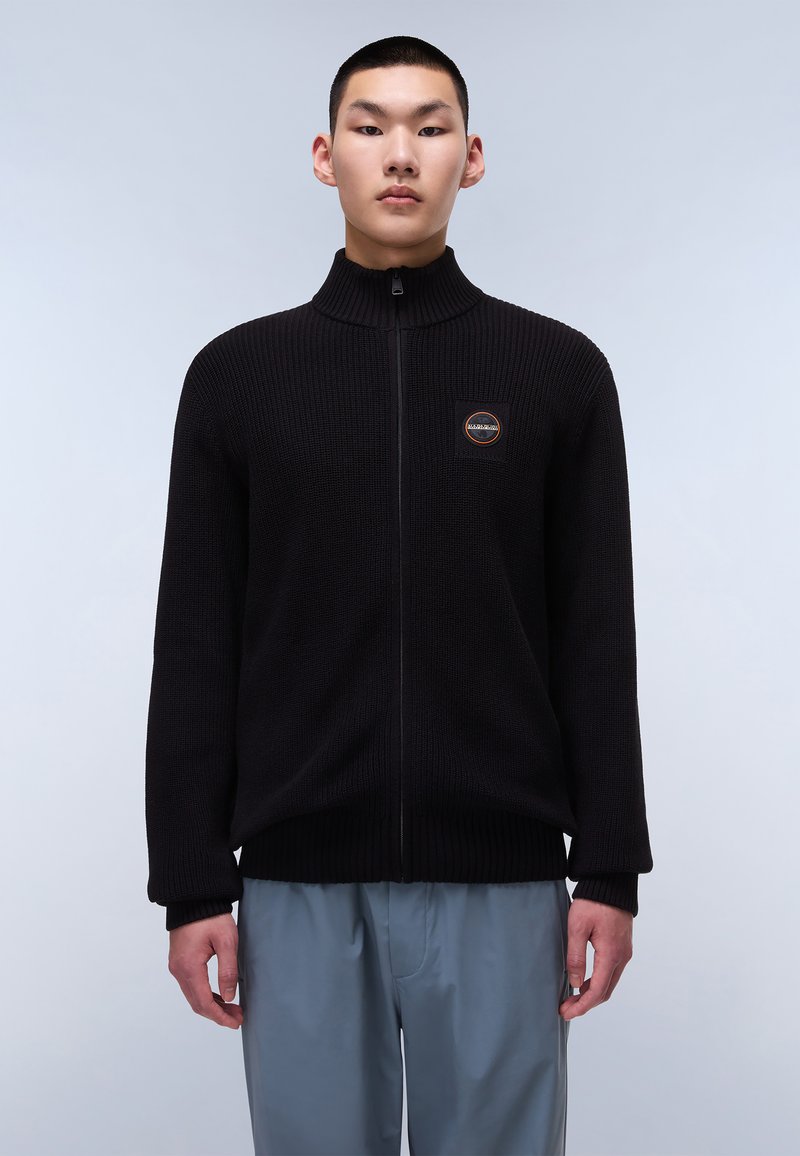 Sweater in maglia nera con zip, colletto alto, texture a coste e una patch con logo sul petto. Modello che indossa pantaloni grigi chiari.