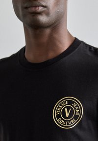 Μαύρη βαμβακερή μπλούζα με κοντό μανίκι, διαθέτει στρογγυλή χρυσή λογότυπη με το κείμενο "VERSACE JEANS COUTURE". Κλασικό λαιμόκοψη.