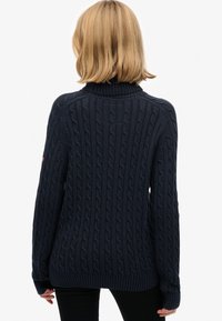 Persona con cabello rubio hasta los hombros, vistiendo un suéter de punto en azul marino y pantalones negros, vista desde atrás contra un fondo blanco.