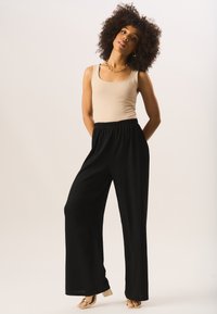 Black, wide-leg pants with a textured finish and elastic waistband, paired with a beige tank top and gold-heeled sandals.