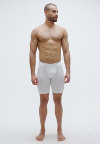 Άσπρα, εφαρμοστά μποξέρκι φτιαγμένα από λείο, ελαστικό ύφασμα με φαρδύ ελαστικό στη μέση και περιγραμματικό ποδογέφυρο, μοντέλο στέκεται ξυπόλυτο μπροστά σε απλό φόντο.