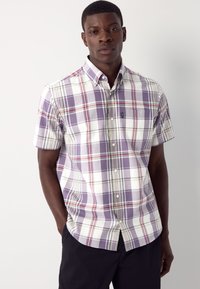 Mees, kes kannab lühikeste varrukatega triiksärki lilla, valge, punase ja rohelise ruudulise mustriga, seisab ühevärvilise heleda tausta ees.