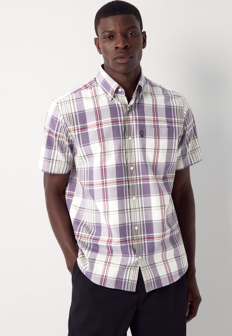 Mees, kes kannab lühikeste varrukatega triiksärki lilla, valge, punase ja rohelise ruudulise mustriga, seisab ühevärvilise heleda tausta ees.