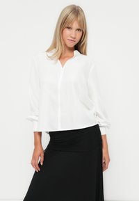 Witte, losse blouse met een kraag, lange mouwen en een knopenrij aan de voorkant; gecombineerd met een zwarte, stretchy rok tot de enkel.