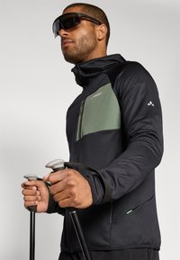 Schwarze technische Jacke mit grünem Brustpanel, durchgehendem Reißverschluss und hohem Kragen. Das Modell hält Trekkingstöcke und trägt dunkle Sonnenbrillen.
