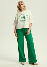 Biały skrócony t-shirt z zielonymi grafikami i cyframi, zestawiony z zielonymi spodniami typu wide-leg oraz jasnobrązowymi trampkami w paski.