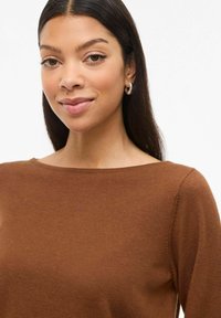Pull en maille marron avec un col rond, texture lisse et silhouette ajustée. Le modèle a les cheveux raides et foncés ainsi que des boucles d'oreilles en argent.