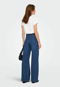 Femme vue de dos portant un haut blanc à manches courtes et un jean bleu à jambes larges, tenant un sac à main noir sur un fond uni clair.