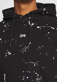 Sweat à capuche noir avec motif de éclaboussures de peinture blanche et petit "274" blanc brodé sur la poitrine, porté avec la capuche relevée et des cordons noirs visibles.