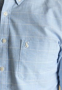 Svetlomodrá kockovaná košeľa s bielymi gombíkmi a náprsníkom s malým bielym vyšívaným logom klokana.