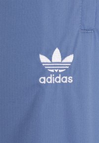Modrá tkanina se strukturovaným vzorem; obsahuje bílé logo Adidas a design tří pruhů na spodní části loga.