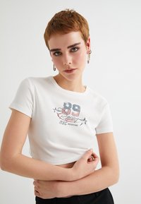 Camiseta blanca corta con mangas cortas, que presenta un diseño gráfico colorido con estrellas y texto, hecha de suave material de algodón.