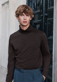 Brun långärmad polotröja i mjukt tyg, kombinerad med blå byxor. Bakgrunden har mörka dörrar och en ljus vägg.