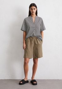 Chemise à manches courtes, boutonnée, avec un motif géométrique noir et blanc, associée à un short en olive avec cordon de serrage et des sandales noires.