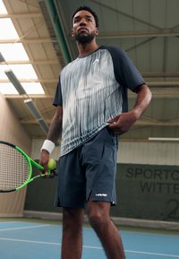 Sportoutfit mit einem kurzärmeligen, grau-weißen, gemusterten Shirt mit blauen Akzenten und dunkelblauen Shorts, während er einen grünen Tennisschläger und einen Ball hält.