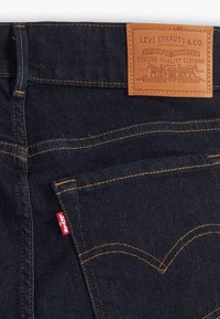 Mörkblå denimjeans med orange sömmar, med en läderlapp med varumärkeslogotyp och en liten röd tagg på sidan.