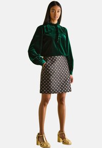 Grüne Samtbluse mit Rüschendetails und langen Ärmeln, kombiniert mit einem schwarzen Rock mit mehrfarbigem Muster und goldenen Blockabsatzschuhen.