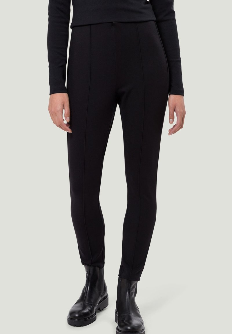 Pantalon noir ajusté avec une texture lisse, doté de détails de coutures à l'avant et d'une taille haute, associé à des bottines noires montant jusqu'à la cheville.