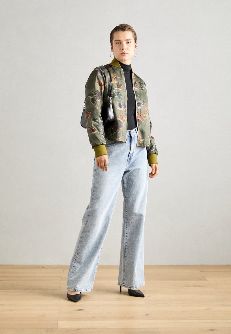 Veste bomber à fleurs en olive et vert, assortie à un jean en denim bleu clair taille haute et des escarpins noirs à bout pointu. Petit sac à main noir en accessoire.