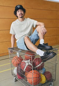 Gray Jordan logo t-shirt, denim shorts, black shoes with contrasting stitching, sitting on a wire cart filled with various basketballs.