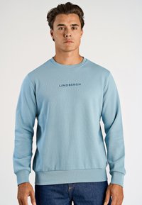 Ljusblå sweatshirt med rund hals, ribbade ärmslut och nedre fåll; har "LINDBERGH"-logotypen i mörkblått över bröstet.