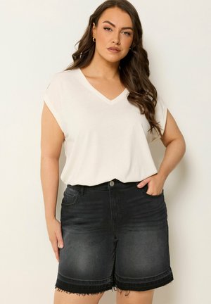 Kvinde med langt brunt hår iført en hvid V-hals T-shirt og sorte denimshorts med frynset kant, stående mod en ensfarvet baggrund.