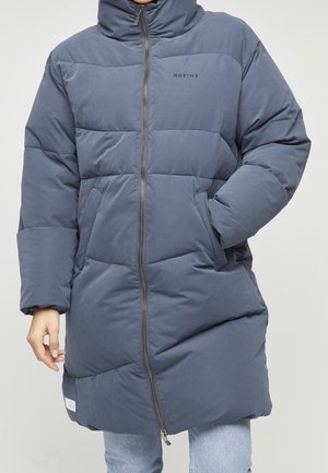 Personne portant un manteau d'hiver long matelassé bleu-gris avec une fermeture éclair à l'avant et des poches latérales, une main dans la poche, l'autre détendue.
