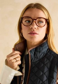 Brūnas bruņurupuču četras brillēm, tumši zils kvilts veste ar zamšādas apkakli un balta blūze ar izšūtiem rakstiem. Zelta gredzens uz rokas.